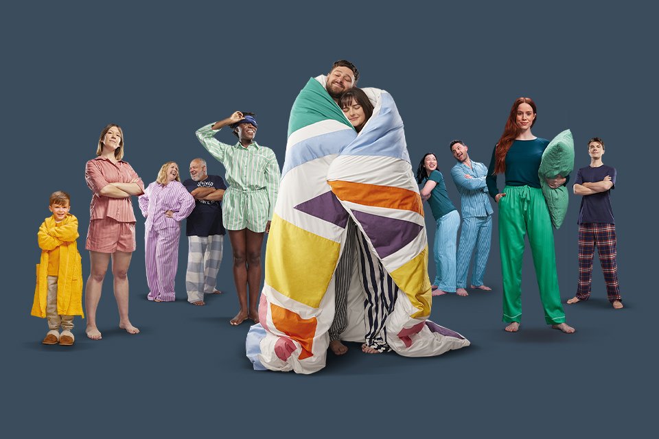 A couple standing and snuggling in a Silentnight duvet with other people in the background.