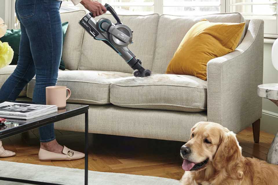 A woman using Vax ONEPWR Blade 3 Dual Pet Cordless Vacuum Cleaner on a sofa while a dog sits next to it.