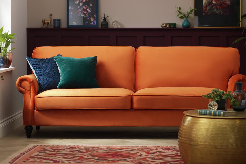 An orange 3 seater fabric sofa bed with green and blue velvet scatter cusions.