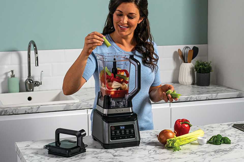 A woman putting chopped vegetables in a Ninja food processor placed on a kitchen countertop.