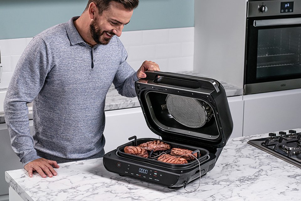 A man in a grey sweatshirt looking at cooked steaks on a Ninja health grill.