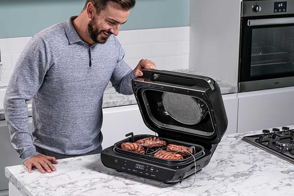 A man in a grey sweatshirt looking at cooked steaks on a Ninja health grill.