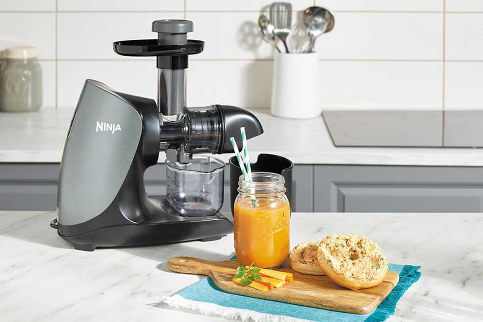 A Ninja juicer with a jar of juice, 2 slices of bread and chopped carrots on a wooden serving board.