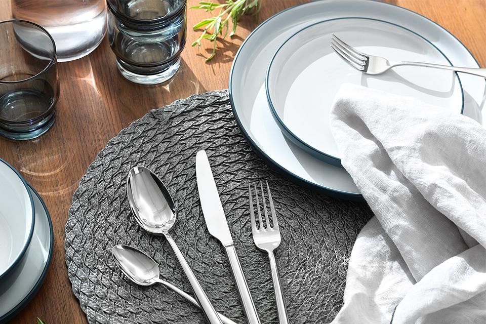 A stainless cutlery set and a white dinner set placed on a grey table mat.