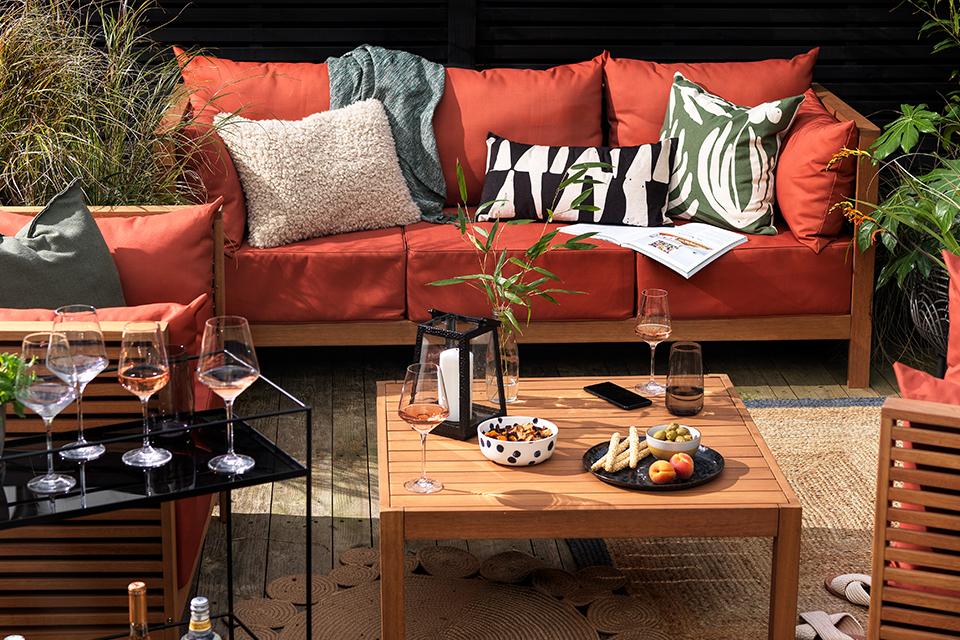 A wooden framed brick red coloured outdoor funiture set on a patio with wine glasses, candle and tableware on the coffee table.