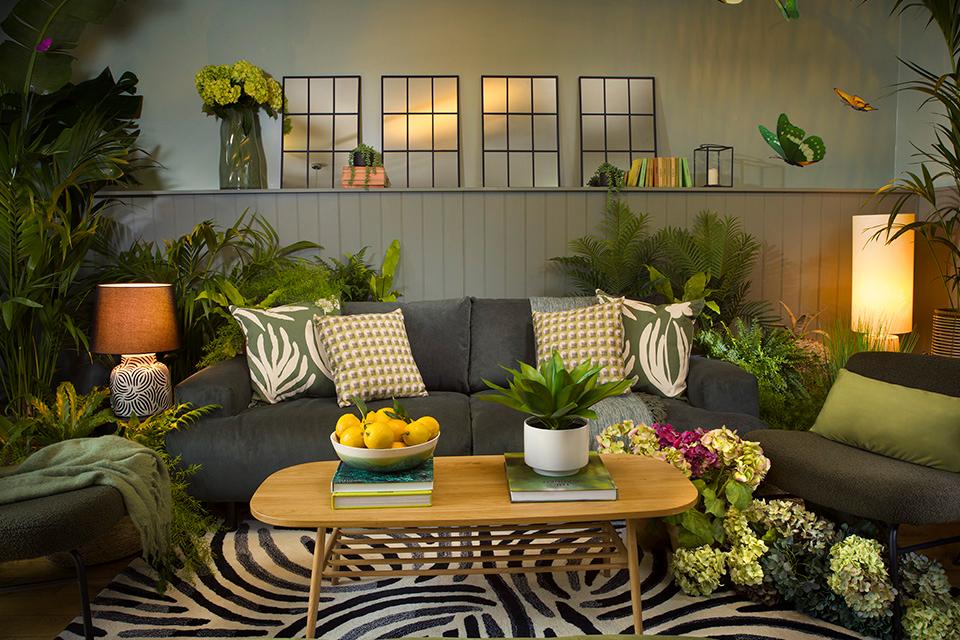 A nature inspired room displaying multiple indoor plants, a grey sofa and leaf print cushions.