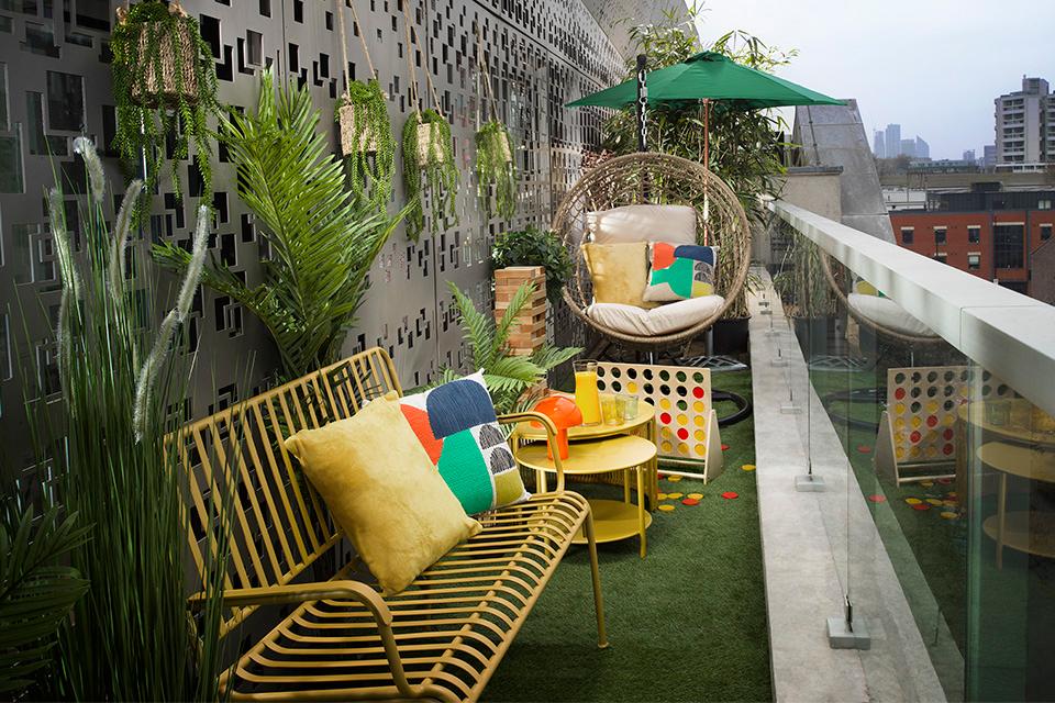 A playroom inspired balcony with a metal garden bench in yellow, multi-coloured cushions, an egg chair and hanging planters with succulents.