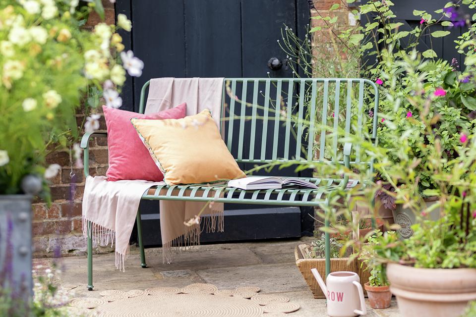 A green garden bench decorated with cushions and a throw.