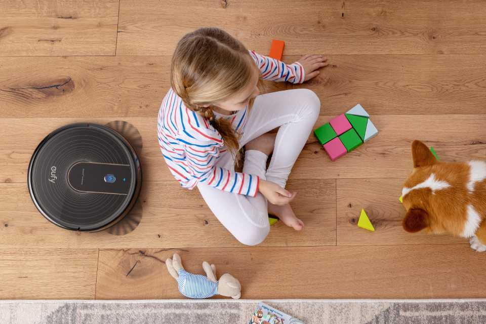 eufy RoboVac 15C quietly cleaning the  floor next to a girl playing with a dog.