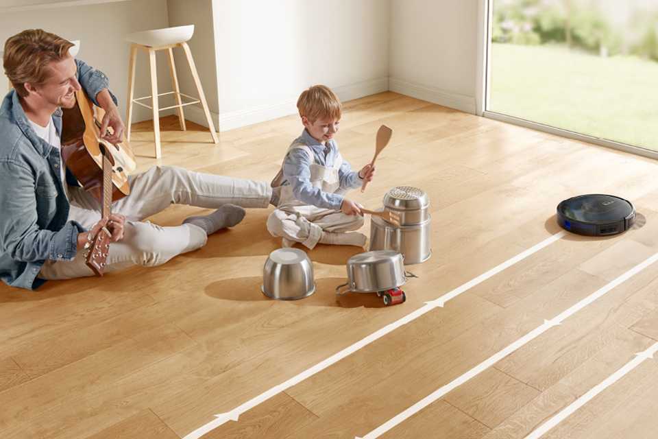 eufy RoboVac G30 navigating and cleaning the floor, next to a child and his father playing music with utensils and guitar.