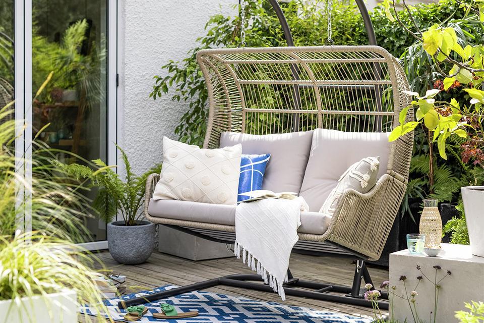 A Habitat double steel garden hanging chair in beige with cushions and a throw on it.