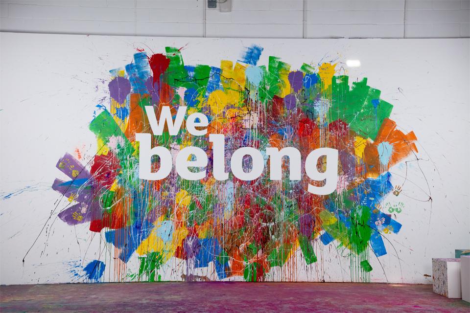 A wall covered in multicoloured paint splashes with the words we belong.