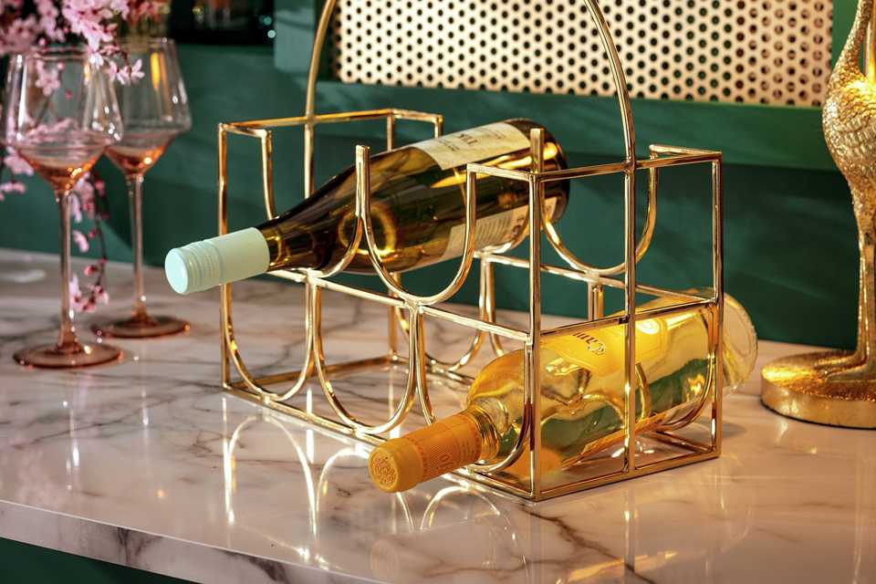 A Habitat stainless steel wine rack for 5 bottles in gold finish placed on a marble counter along with 2 wine glasses.