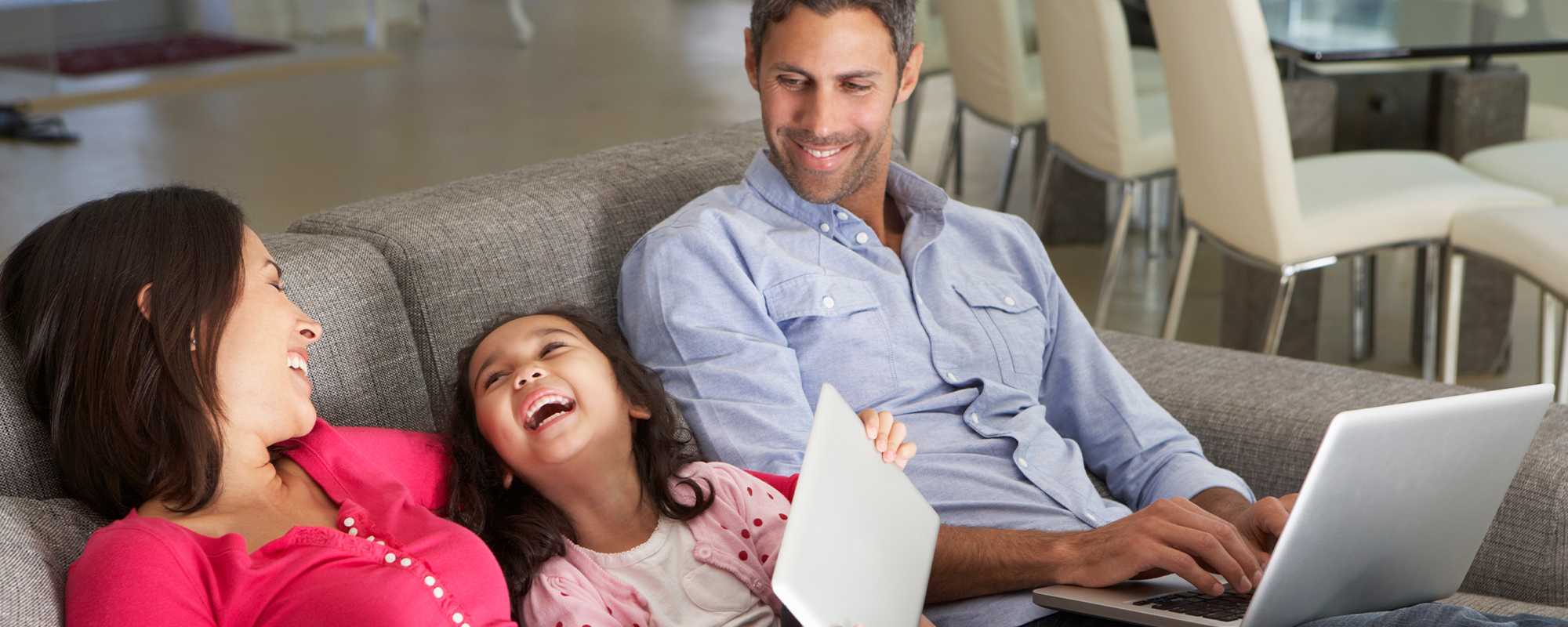 A laughing family sit on a sofa with laptops.