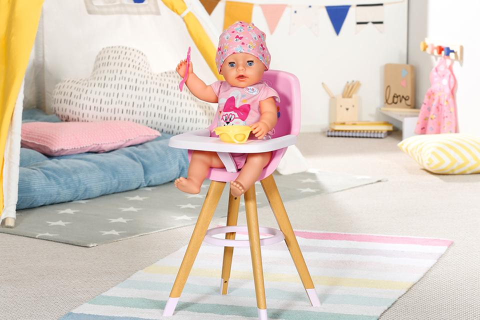 A BABY born doll holding a spoon and a bowl while sitting on a highchair.