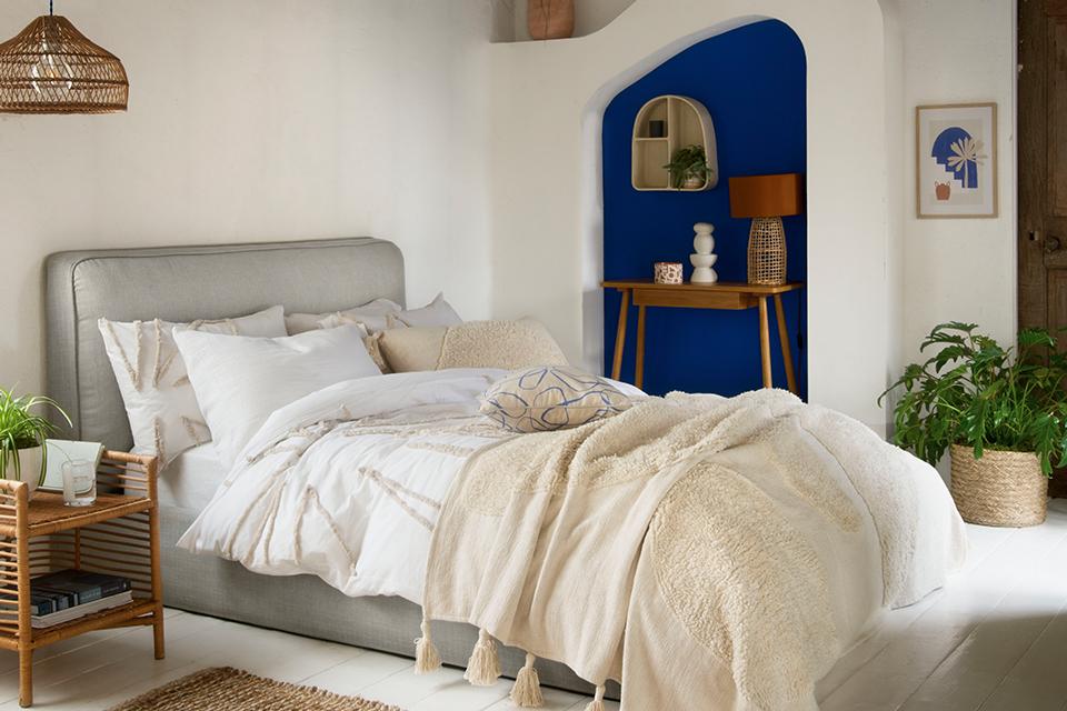 A mediterranean theme bedroom with a grey bed and white bedding, white wall shelving and natural finish furniture. 