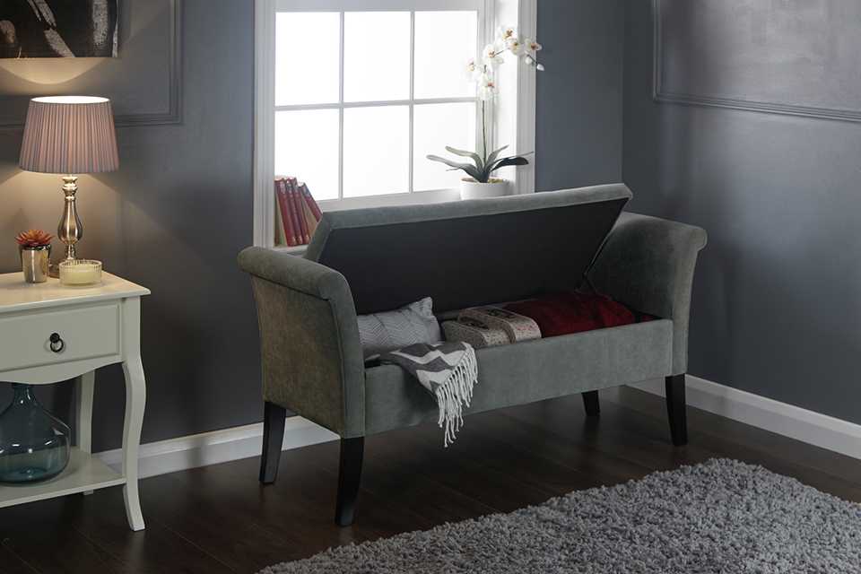 A grey fabric ottoman seat with storage next to a window. 