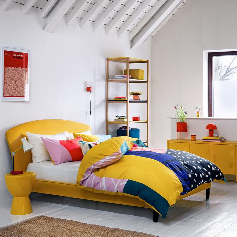A colourful bedroom with a mustard and blue duvet, a side table and storage.
