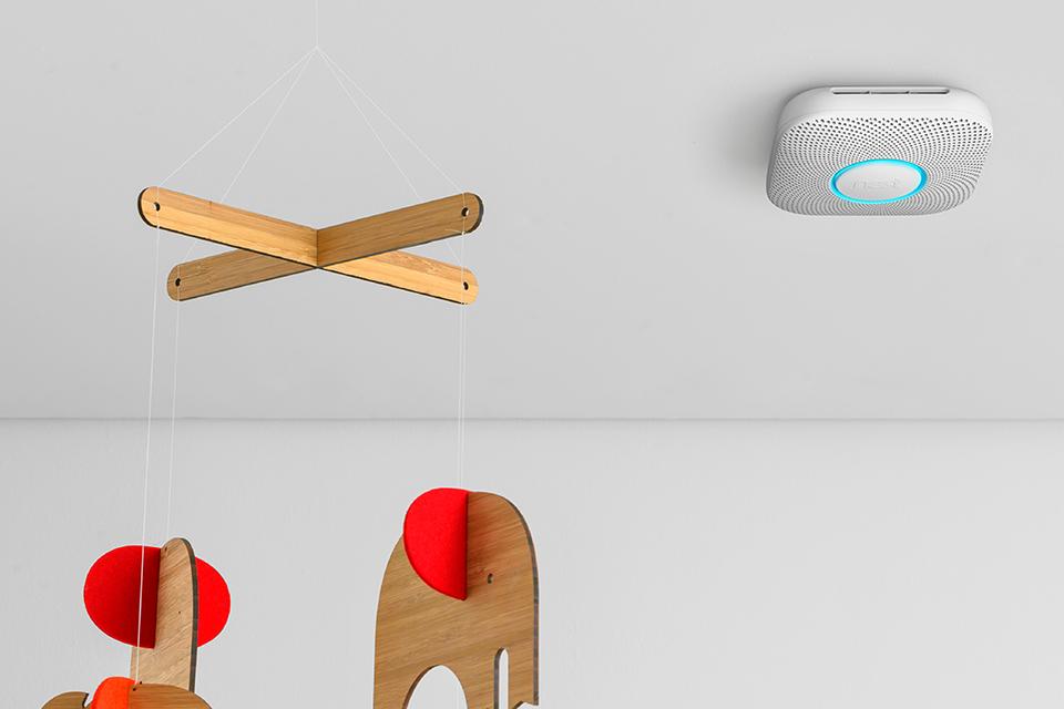 A Google Nest smoke and CO detector installed on the ceiling next to a wooden animal caper mobile.
