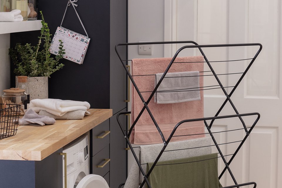 An image of 3 tier indoor clothes airer in black with t-shirts and towels drying on it placed next to a washing machine. 