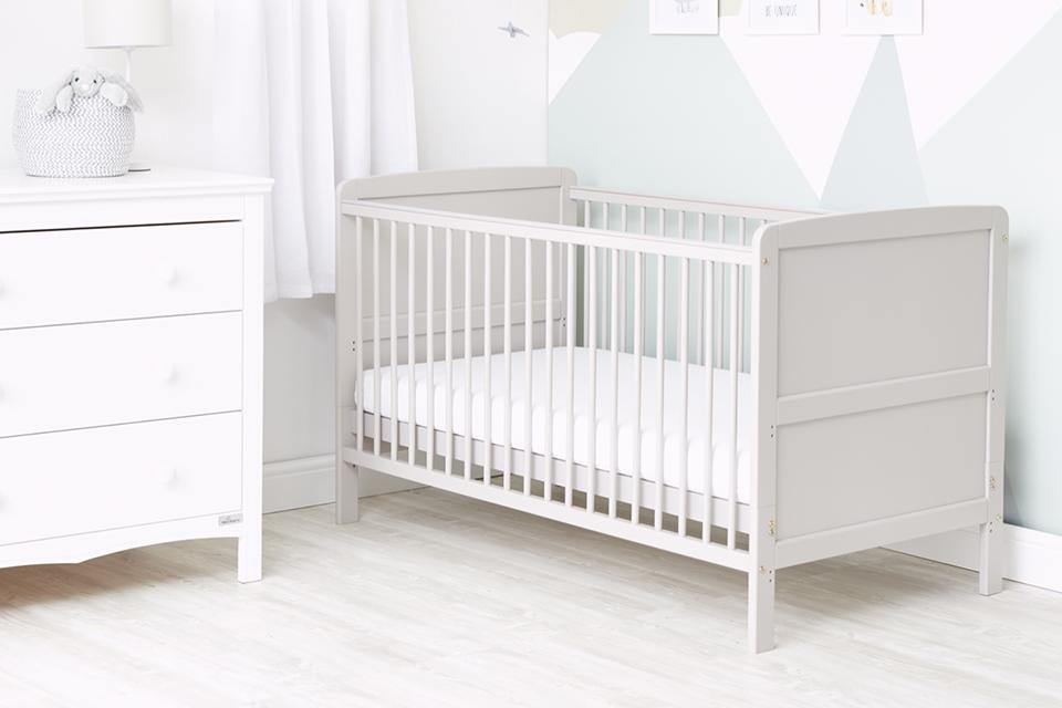 A Baby Elegance baby cot bed with mattress in white placed next to a chest of drawers in a kid's room. 