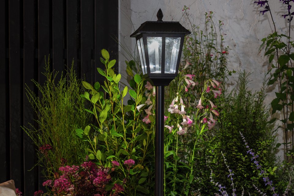 A black Argos Home Metro solar powered lampost in a garden next to some flowers.