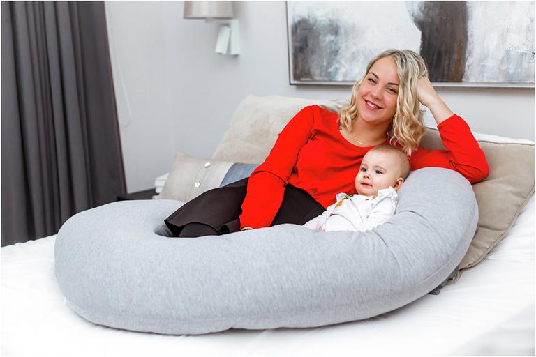 A mother and her baby resting on a bed with a Kolbray c shaped pregnancy pillow in grey colour.