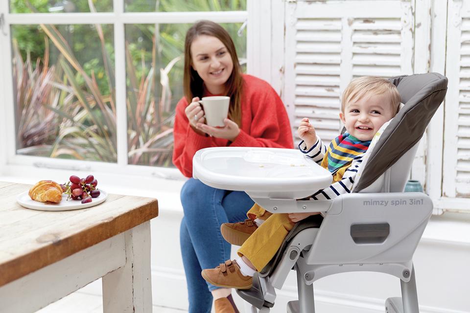 A baby sitting on a Joie Mimzy grey and white highchair near a dining table.