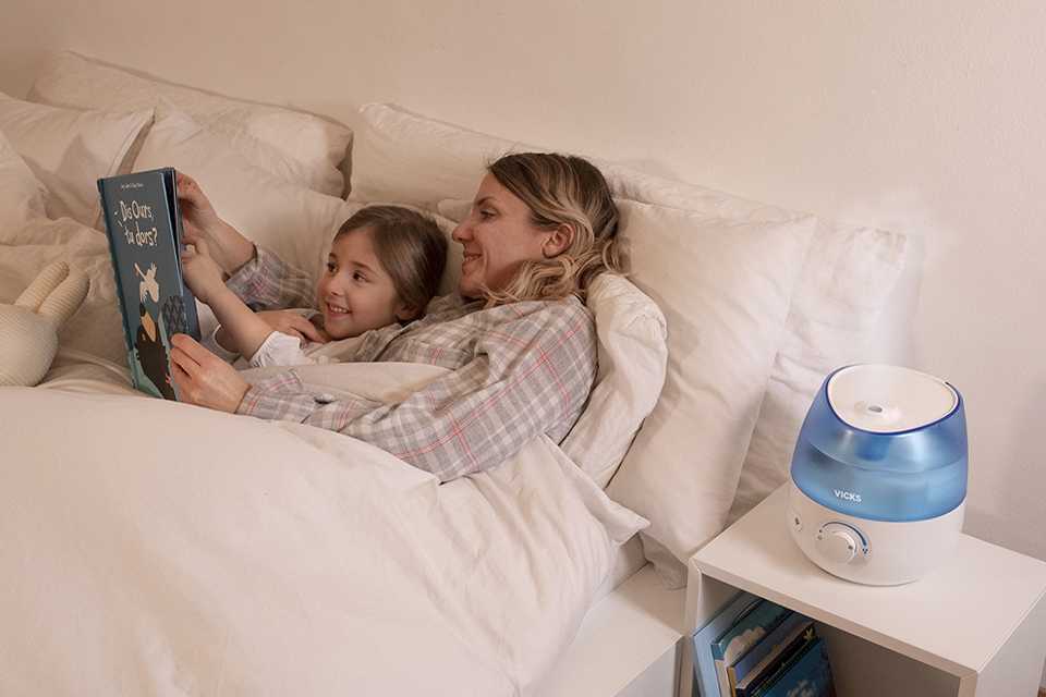 A Vicks humidifier alongside a bed with mother and daughter reading a nighttime book.