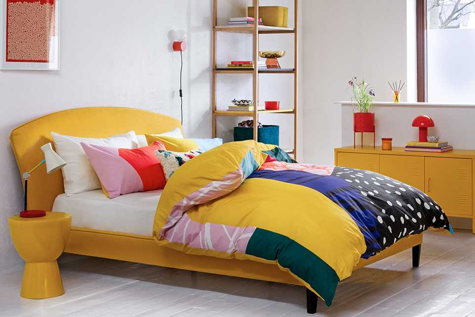A white bedroom with bold and bright coloured furniture including a yellow bed, sidetable and multi-coloured bedding and cushions. 