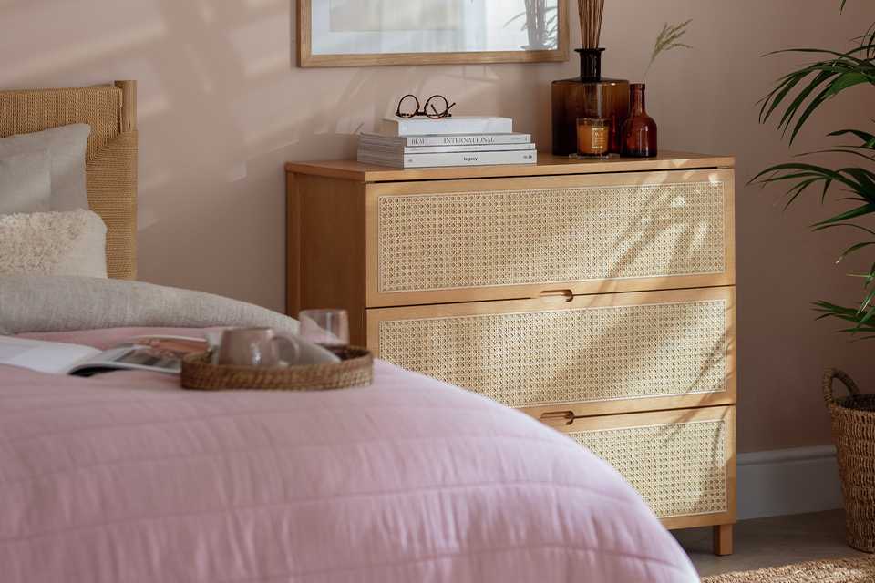 A cane 3 drawer storage chest next to a bed in a pink bedroom.