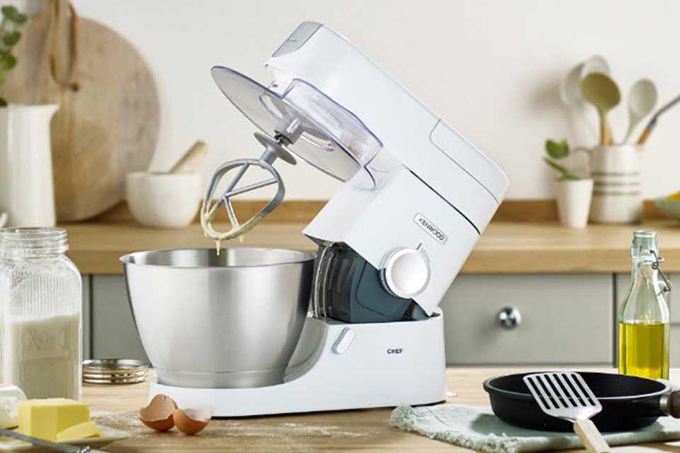 A white Kenwood stand mixer on a cluttered kitchen surfact.