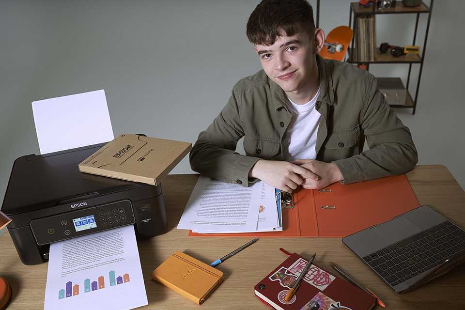 A boy sitting on a desk with an open file folder with documents next to an Epson printer and the ReadyPrint Flex card.