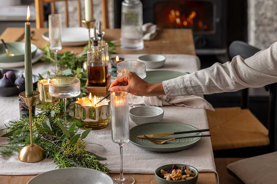 A person lighting a candle placed on a dining table decorated for Christmas with tableware, glassware and cutlery. 