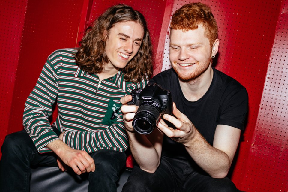 Two friends smiling while looking at pictures on a Canon EOS 2000D camera. 