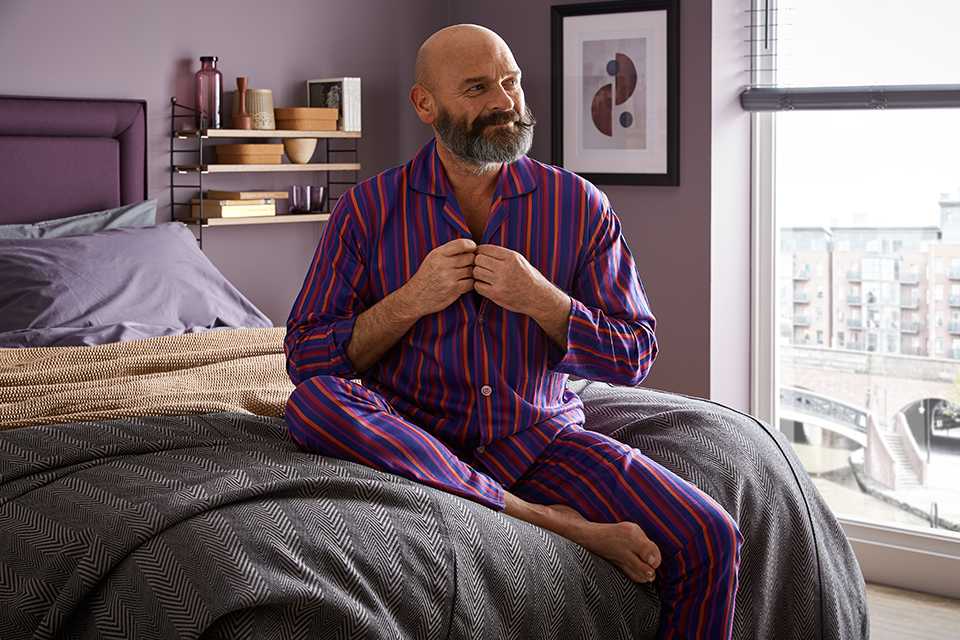 A man in pajamas sitting on a Silentnight bed next to a window.