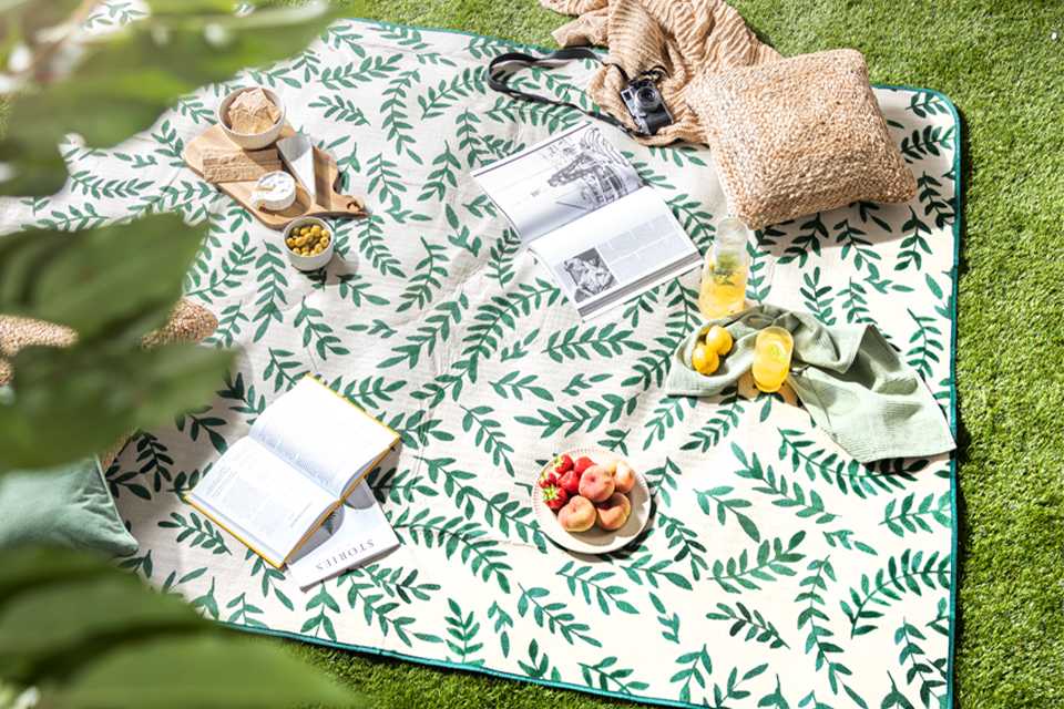 A white and green leaf-print blanket with picnic set up including books, cushion, glassware, and serveware.