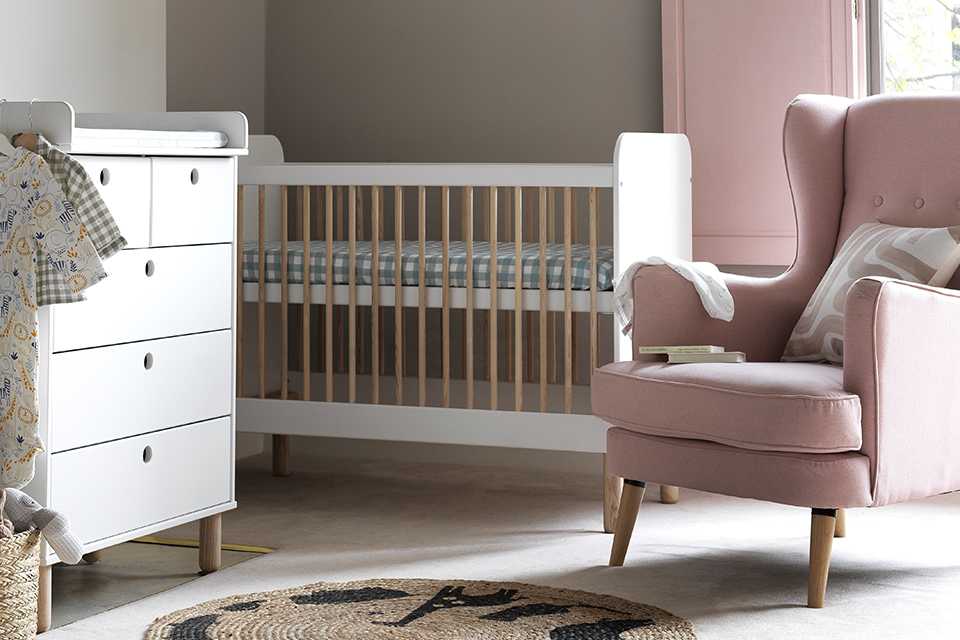 A nursery with white and wood finish cot, chest, and a pink armchair. 