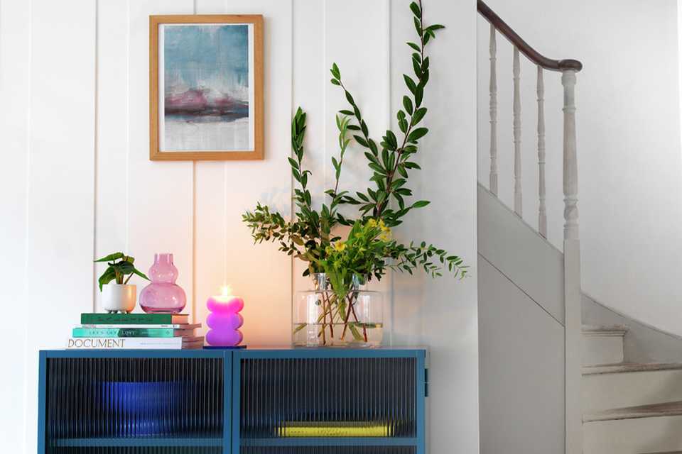 A blue ribbed glass sideboard with an indoor plant, a lilac vase, a glass vase, and a pink candle on it.