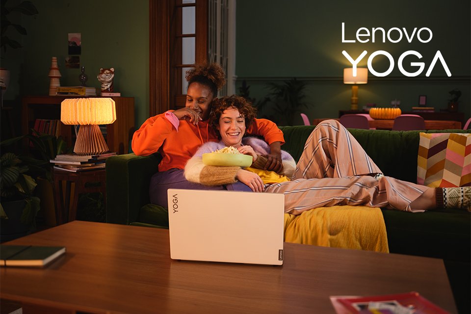 Two smiling women eating popcorn while watching a movie on a Lenovo Yoga laptop.