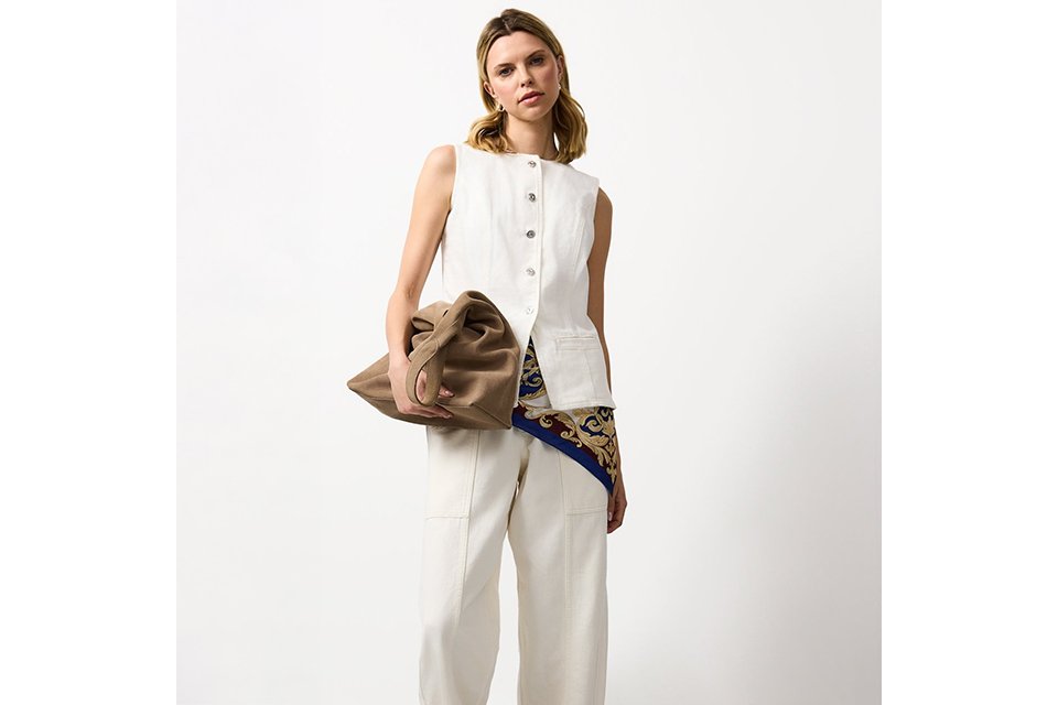 Cream sleeveless top and matching skirt styled with a black cut‑out bucket bag and crossbody strap on a neutral studio background.