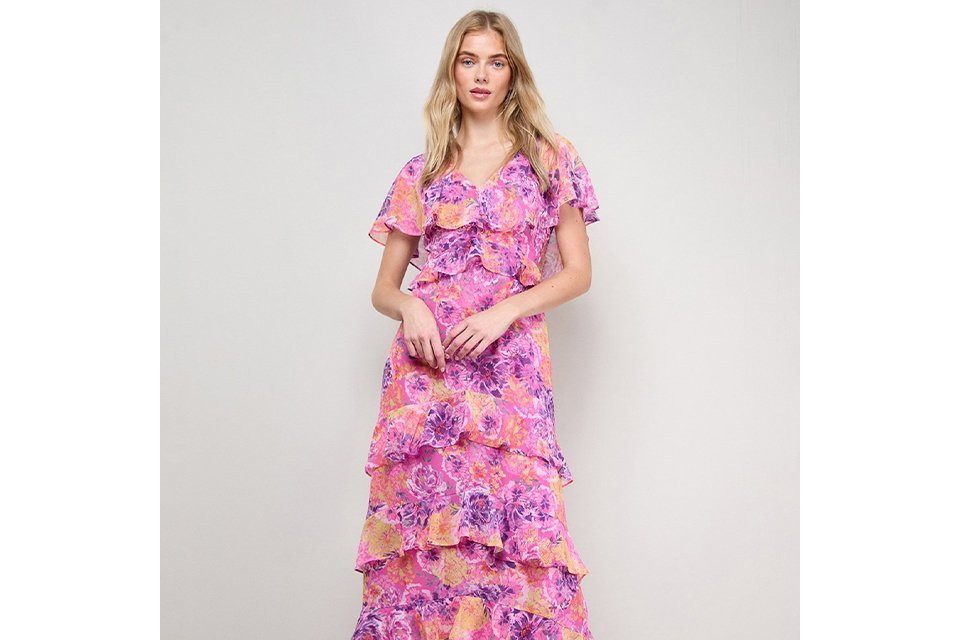 Pink floral tiered midi dress with short flutter sleeves and V‑neckline, styled with neutral heeled sandals on a light studio background.