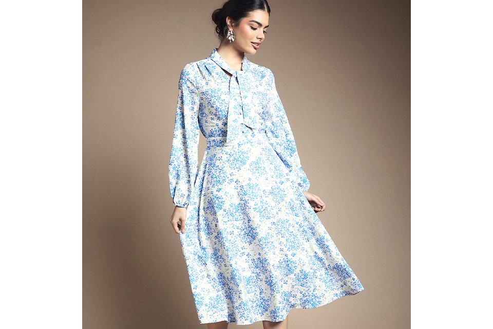 Blue and white floral long‑sleeve midi dress with tie‑neck detail and softly flared skirt on a neutral studio background.