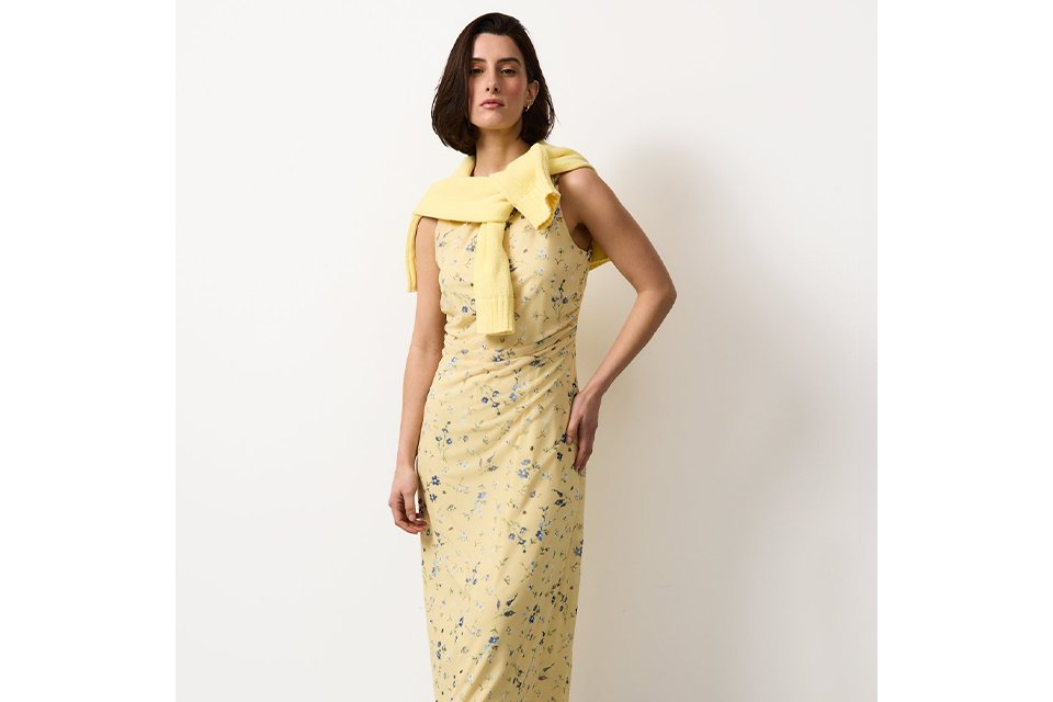 Women wearing a yellow dress with flowers on and a yellow jumper draped over her shoulders. 