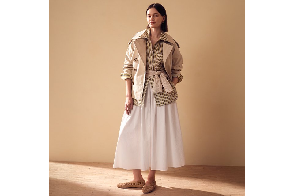 A women wearing a white maxi skirt with a cream striped shirt and cream short trench jacket.