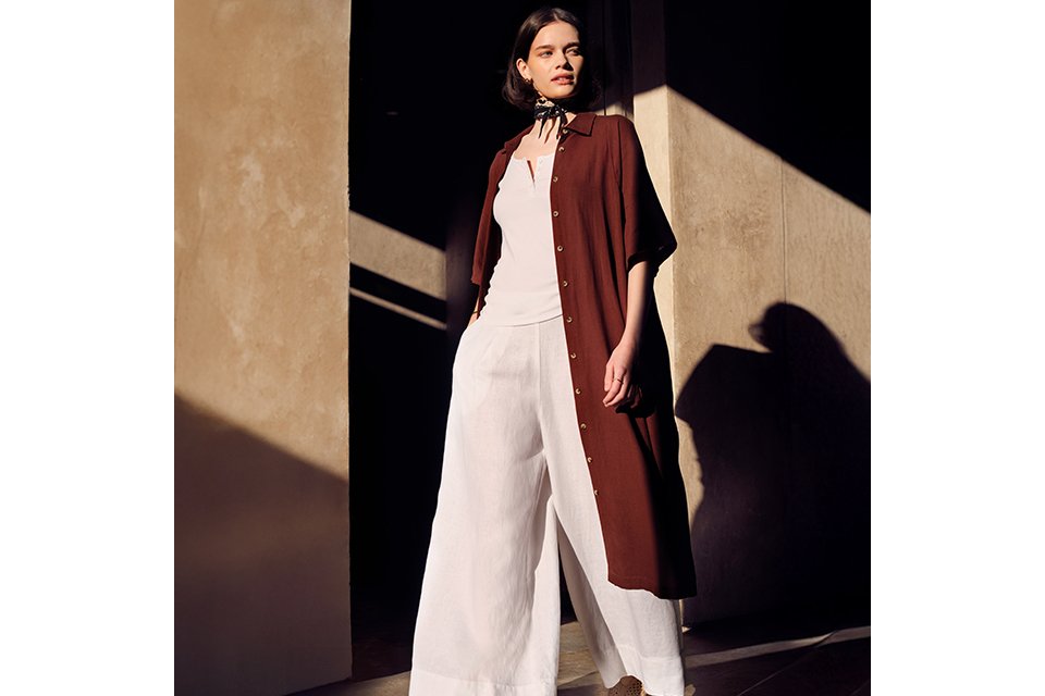 A women wearing white linen trousers and white top with a longline burgundy shirt.