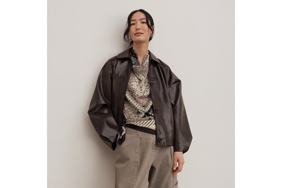 Model wearing a dark leather-look jacket layered over a patterned shirt with neutral trousers for transitional spring dressing.