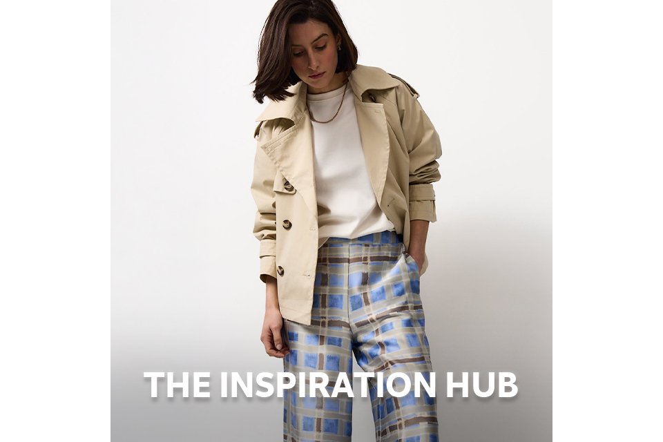 Woman wearing blue and brown check trousers, with neutral t-shirt and stone cropped trench jacket.
