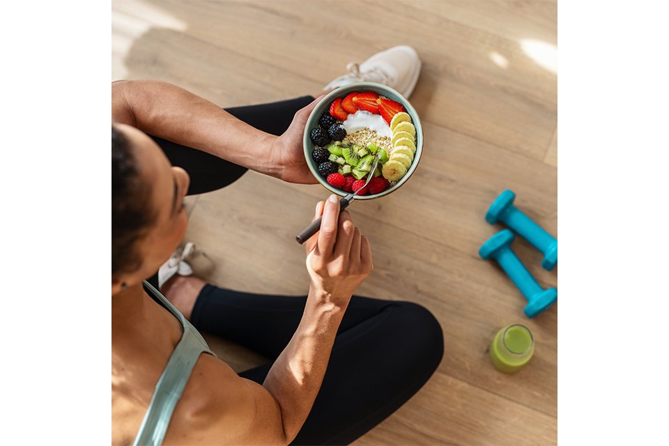 Person sitting on a wooden floor holding a bowl of colorful fruit and yogurt, with two blue dumbbells and a green drink bottle nearby.