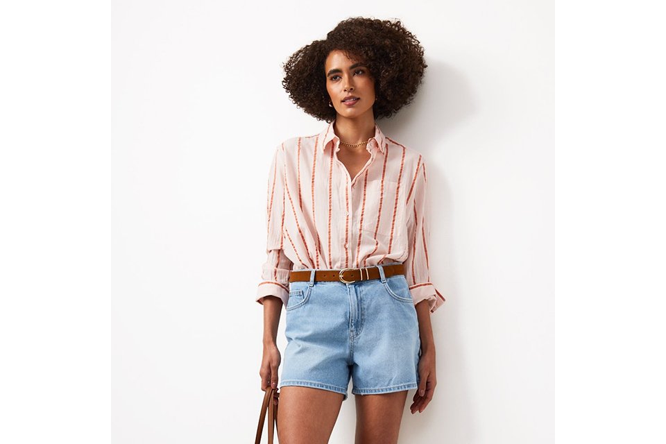 A woman wearing denim shorts, stripey shirt and carrying a straw bag with slip on shoes.